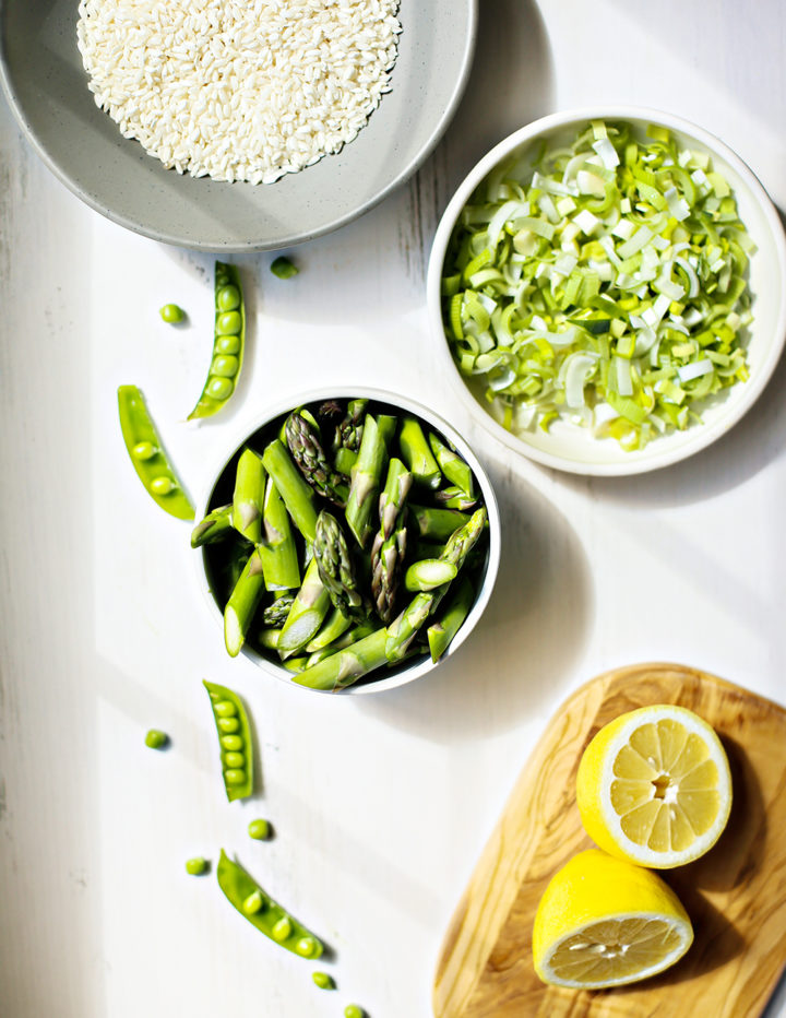 A flat lay of key ingredients for lemon asparagus risotto: a bowl of uncooked arborio rice, a glass of dry white wine, garlic, mascarpone, parmesan cheese, and a bundle of asparagus on a wooden board. Fresh peas and lemons hint at the risotto’s fresh, bright flavors.