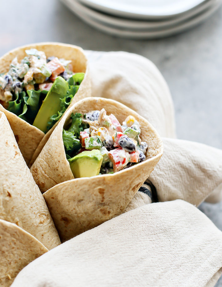 Close-up of a Southwest Chicken Salad Wrap on a light gray surface. The tortilla is tightly wrapped and filled with a creamy chicken salad mixture, black beans, corn, red bell pepper, fresh lettuce, and avocado slices.