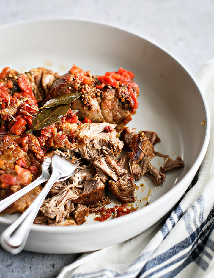 Cooked pork shoulder for pork tacos is shown being shredded with two forks in a shallow white bowl. Juicy chunks of pork are mixed with diced tomatoes and bay leaves, revealing a tender, flavorful texture. A kitchen towel is wrapped around the base of the bowl.