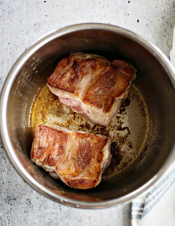 Overhead view of two seared pieces of pork shoulder browning in the bottom of an Instant Pot for a shredded pork taco recipe. The pork is golden brown and resting in rendered juices, showing the early stage of cooking. The background includes a white and navy striped towel and a lightly textured surface.
