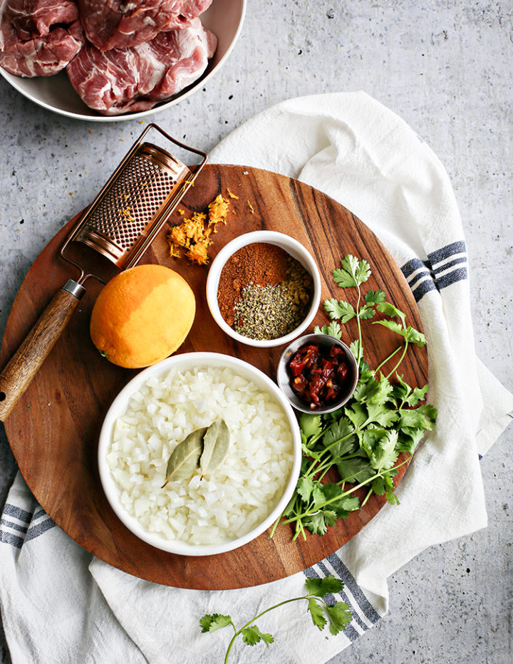 Overhead image of shredded pork taco ingredients arranged on a round wooden cutting board. A bowl of diced white onion with bay leaves, fresh cilantro sprigs, dried spices, chipotle in adobo, orange zest, and a whole orange are styled beside a metal grater and raw pork shoulder in a separate bowl. A white and blue towel adds texture to the bright prep scene.