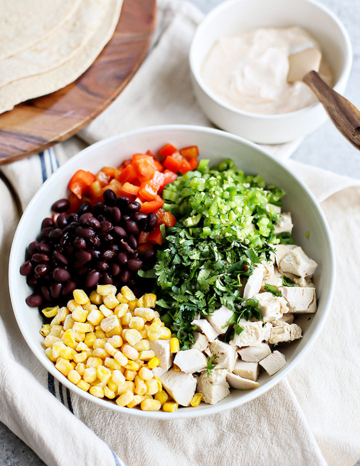 Ingredients in Santa Fe Chicken Salad Wrap – A large white bowl contains diced cooked chicken, black beans, corn, red bell pepper, chopped jalapeño, and cilantro, arranged in colorful sections. A smaller bowl of creamy dressing and a stack of flour tortillas sit nearby on a wooden cutting board.