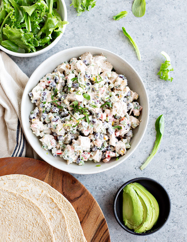 Overhead view of ingredients for assembling Southwest Chicken Salad Wraps, arranged on a light gray background. A white bowl holds the creamy chicken, black bean, corn, and veggie mixture, while a separate bowl contains fresh lettuce. A plate of soft flour tortillas sits nearby, along with a small bowl of sliced avocado, ready for wrapping