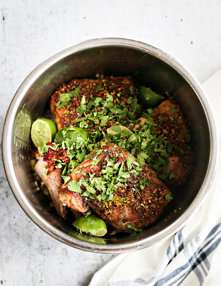 Top-down view of pork shoulder inside an Instant Pot, topped with chopped cilantro, chili flakes, orange zest, and surrounded by halved limes. The seasoned pork is ready for pressure cooking. The scene is bright with a white and blue striped towel nearby.