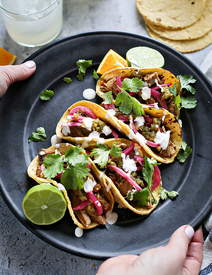 woman holding a black plate with shredded pork tacos on it and a margarita in the background