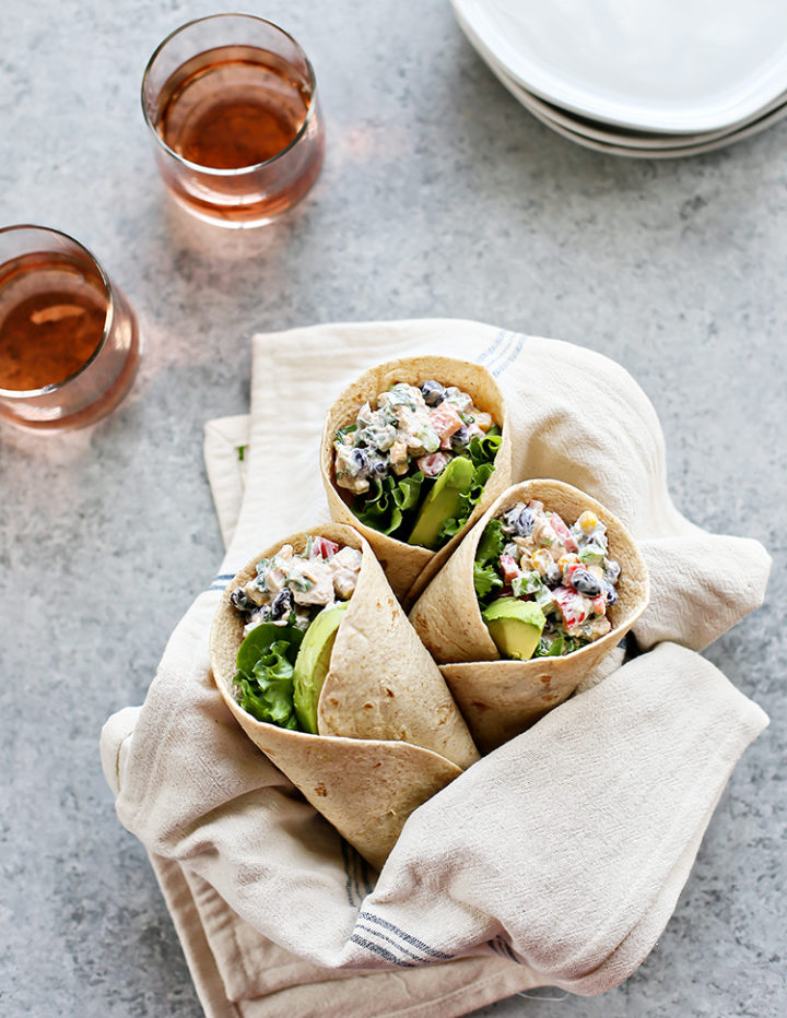 Easy Chicken Salad Wrap – Three Southwest Chicken Salad Wraps, cut in half, are nestled in a linen-lined basket. The wraps are filled with a creamy chicken salad mixture, black beans, corn, red bell pepper, and avocado slices. Two glasses of rosé and a stack of plates are in the softly blurred background.