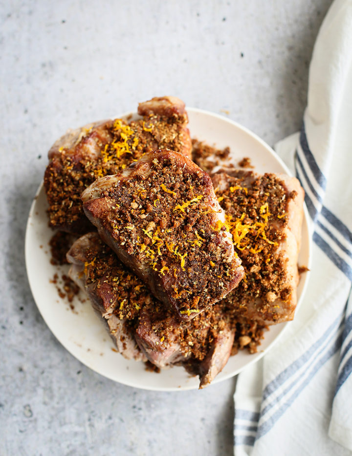 Overhead shot of seasoned pork shoulder chunks piled on a white plate. The meat is coated in a rich, Pulled pork taco seasoning and sprinkled with orange zest. A white and blue kitchen towel lines the background, adding a cozy, homemade feel.