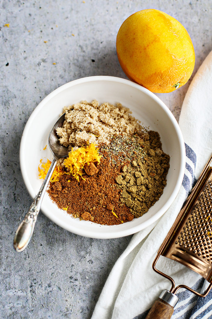 A white bowl holds a variety seasonings for shredded port tacos, along with a small mound of orange zest. A spoon rests inside the bowl, and a whole orange and grater are nearby on a textured surface, suggesting a vibrant and aromatic spice blend.
