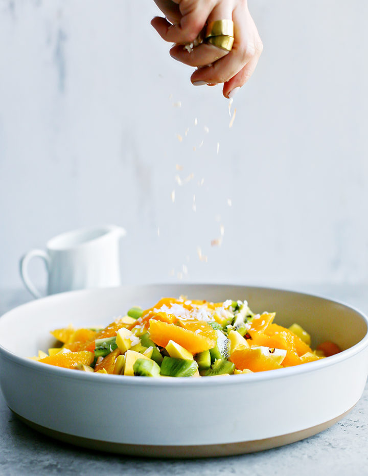 a photo of a woman sprinkling toasted coconut on top of a bowl of tropical fruit salad