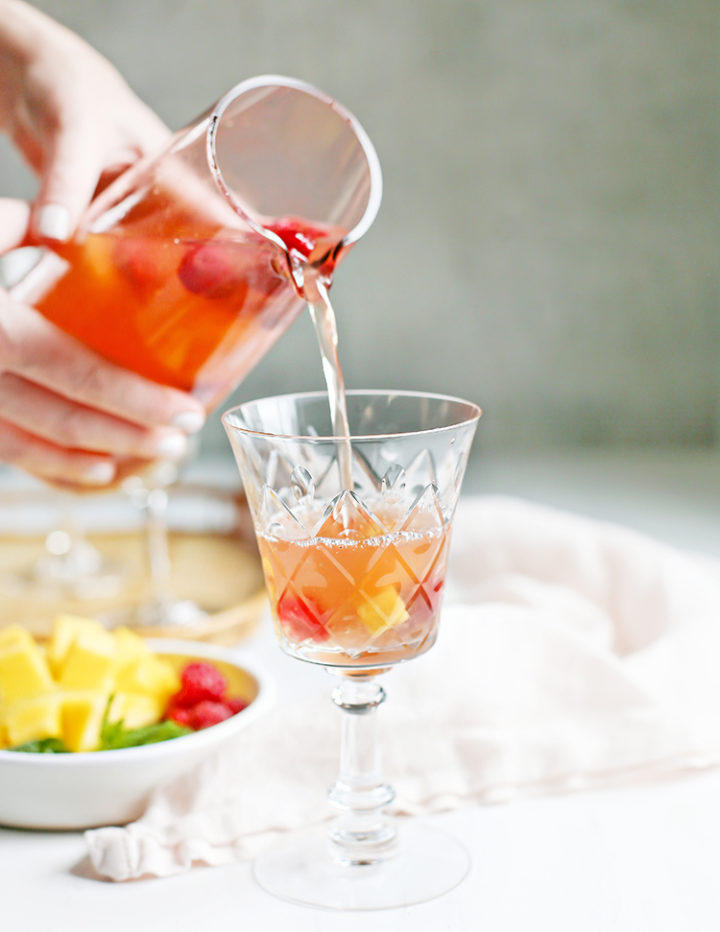 photo of a woman pouring a pitcher of rosé sangria into a glass with raspberries and mango