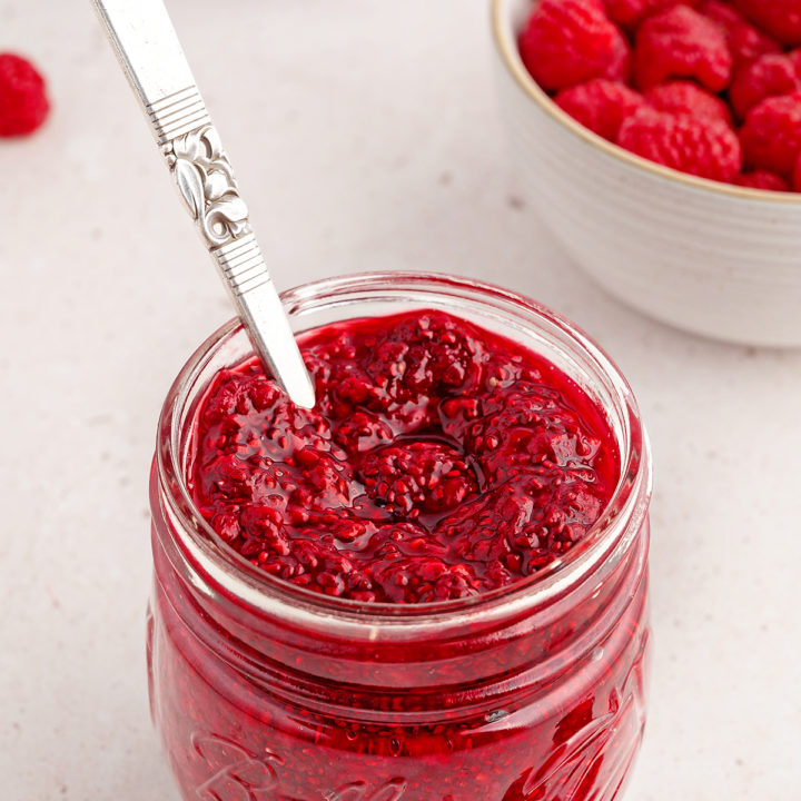 photo of chia jam for a recipe to make raspberry jam in a jam jar with a spoon and bowl of fresh raspberries