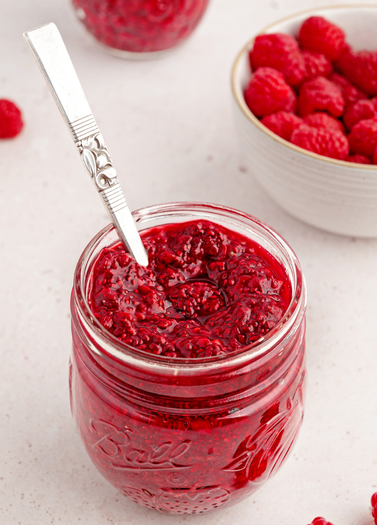 photo of chia jam for a recipe to make raspberry jam in a jam jar with a spoon and bowl of fresh raspberries