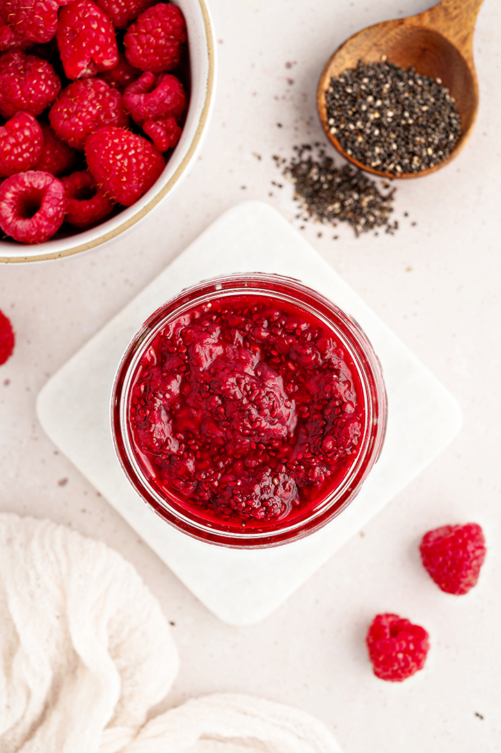 overhead photo of a jar of raspberry jam with chia seeds and fresh raspberries