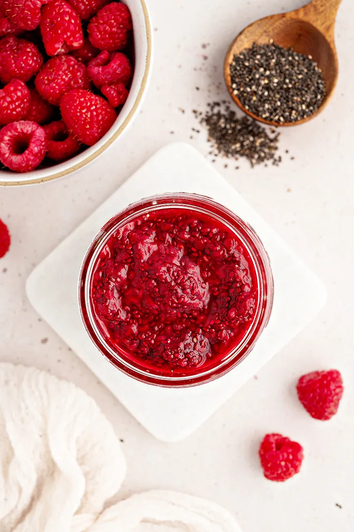 overhead photo of a jar of raspberry jam with chia seeds and fresh raspberries