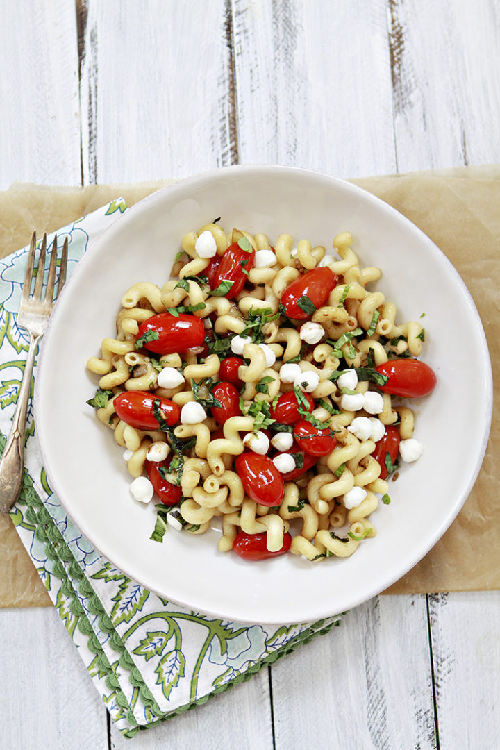 overhead photo of a white pasta bowl with pasta caprese and a basil garnish on a white wooden table