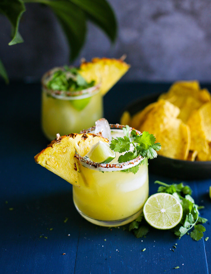 photo of two pineapple margaritas on a dark blue background with a bowl of tortilla chips