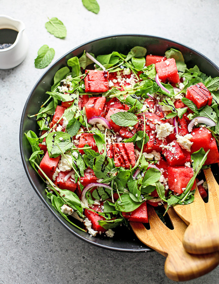 photo of watermelon feta mint salad with balsamic glaze in a black bowl with wooden salad tongs on a wooden table
