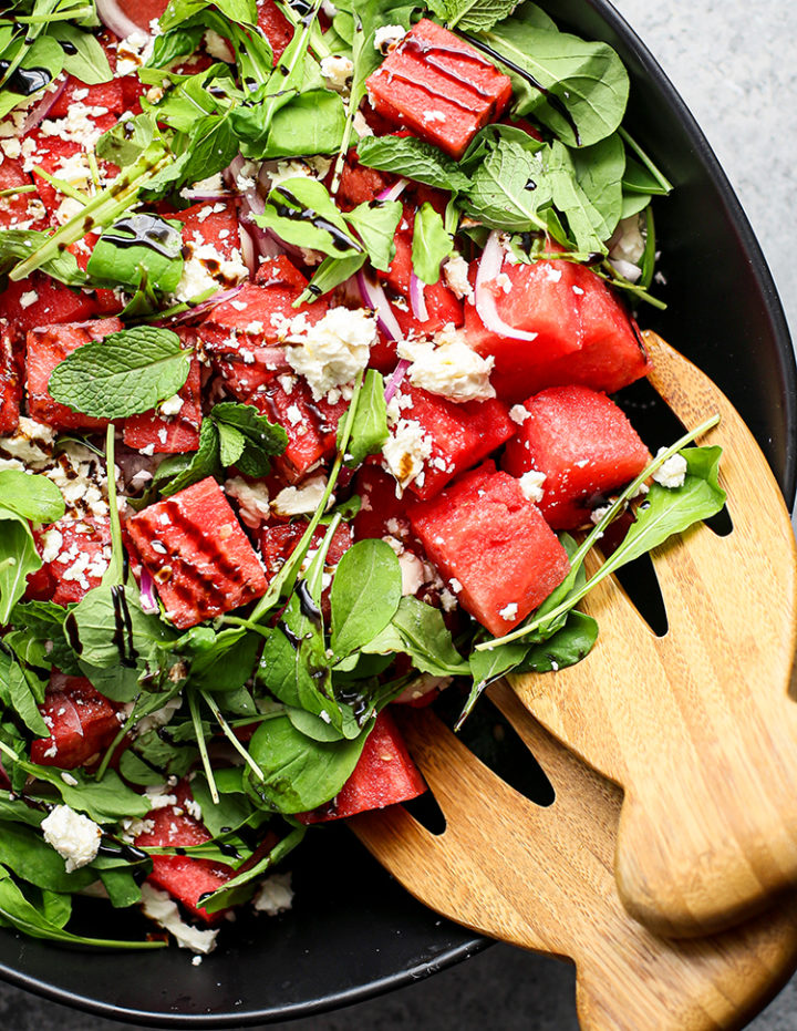 close up photo of watermelon feta mint salad with balsamic glaze in a black bowl with wooden salad tongs
