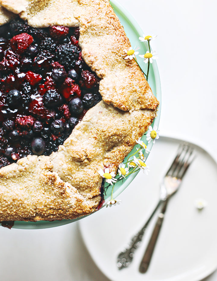 close up photo of a mixed berry galette on a cake stand with plates and forks for serving