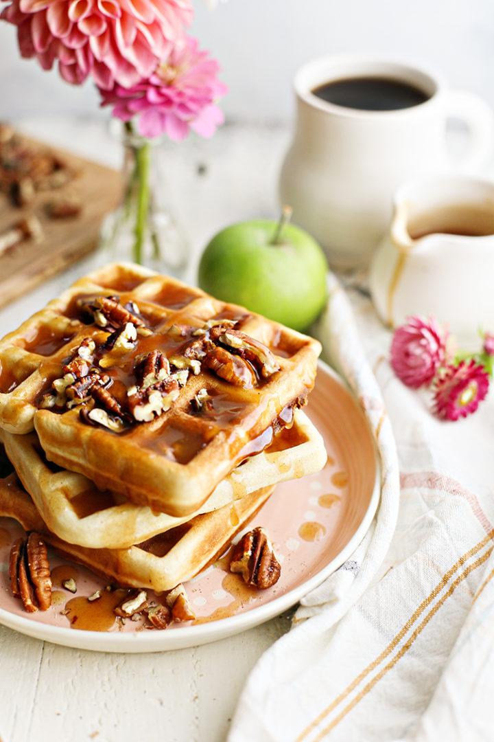 photo of a stack of 3 pecan waffles on a plate set at a breakfast table with a mug of coffee and vase of flowers