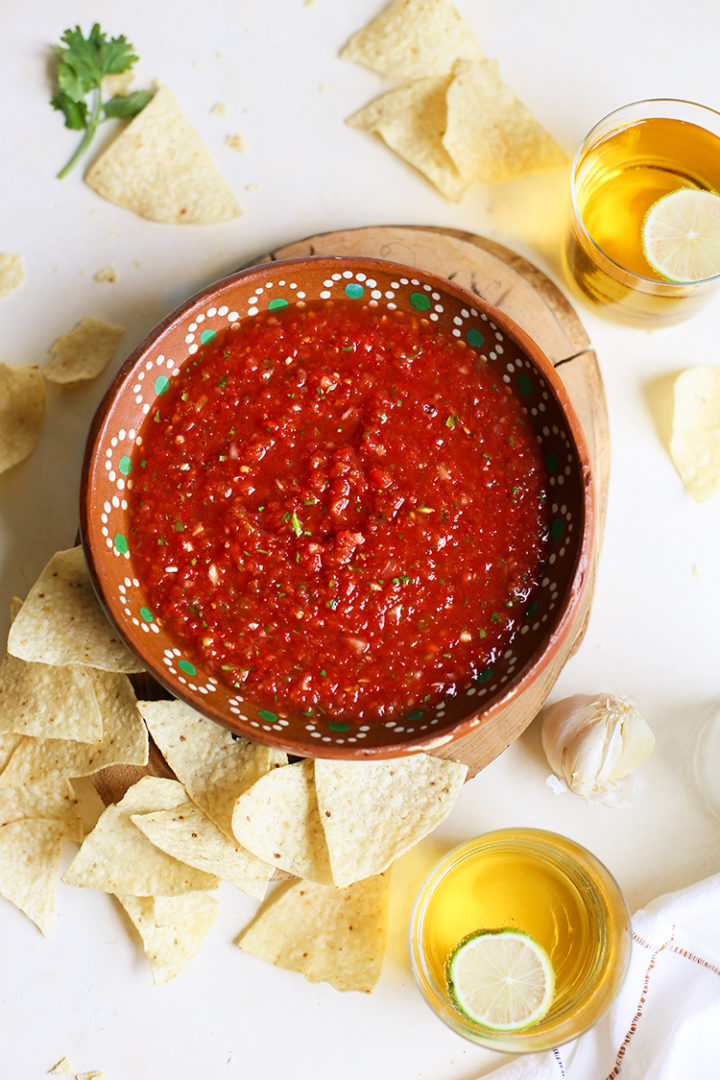 overhead photo of a bowl of salsa restaurant style surrounded by tortilla chips, cilantro, and garlic with two glasses of beer