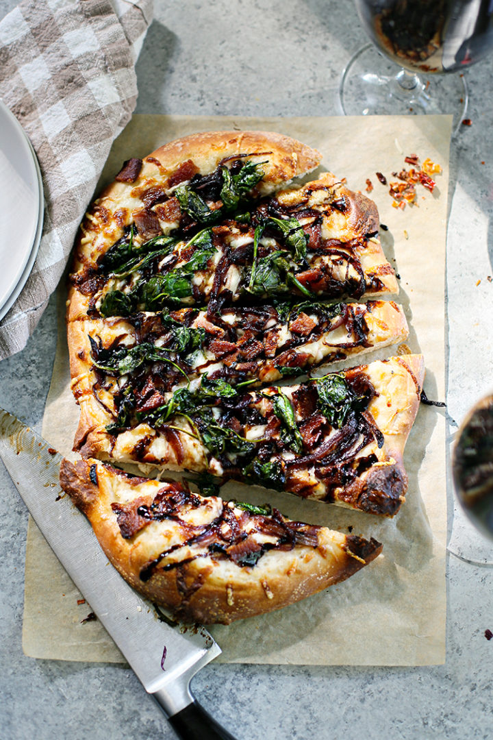 overhead photo of cut slices of Bacon Pizza with Caramelized Onion and Spinach next to a knife and plates