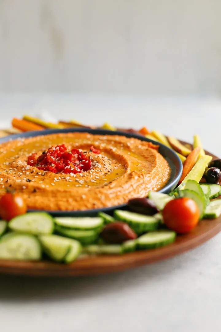 photo of red pepper hummus in a bowl surrounded by vegetables