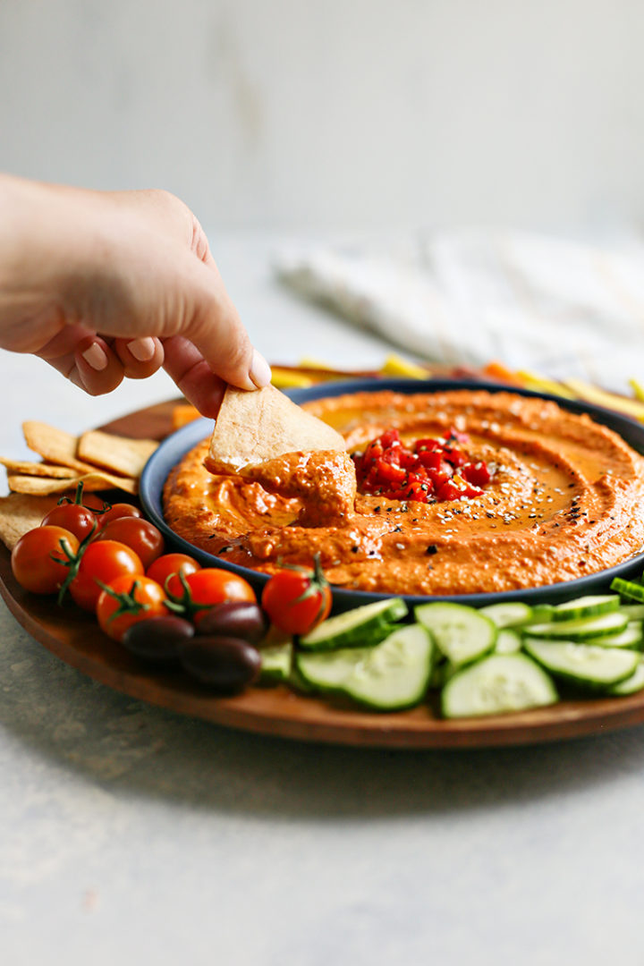 photo of a woman dipping a pita chip in a bowl of red pepper hummus 