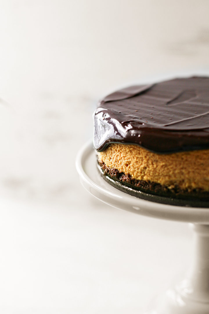 photo of a pumpkin cheesecake with chocolate crust and chocolate ganache topping on a white cake stand