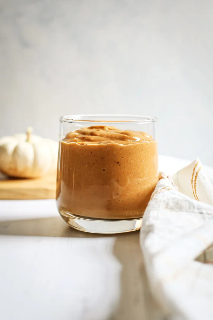 photo of a pumpkin pie smoothie in a cup on a white table with a small pumpkin and plaid dish towel