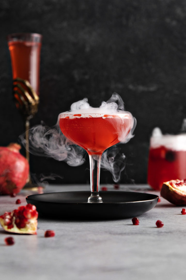 photo of a pomegranate halloween cocktail with dry ice and a fake blood cocktail rim on a black plate with a dark background