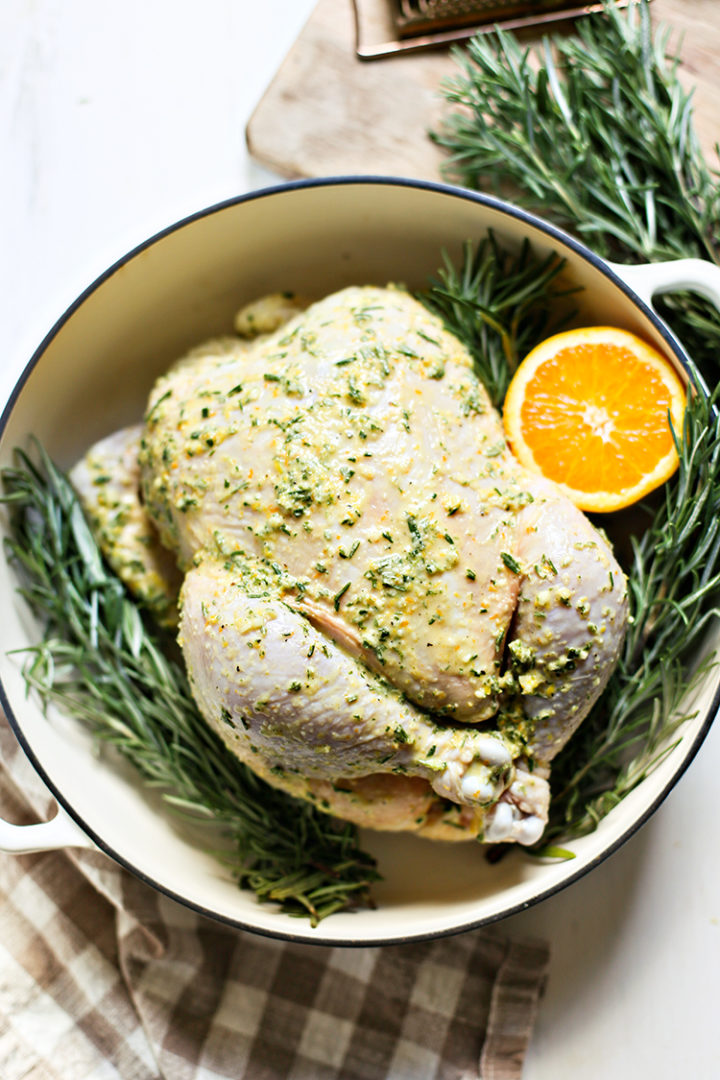 photo of a whole chicken with citrus rosemary marinade, fresh rosemary sprigs, and orange slices in a white cast iron roasting pan ready to be roasted