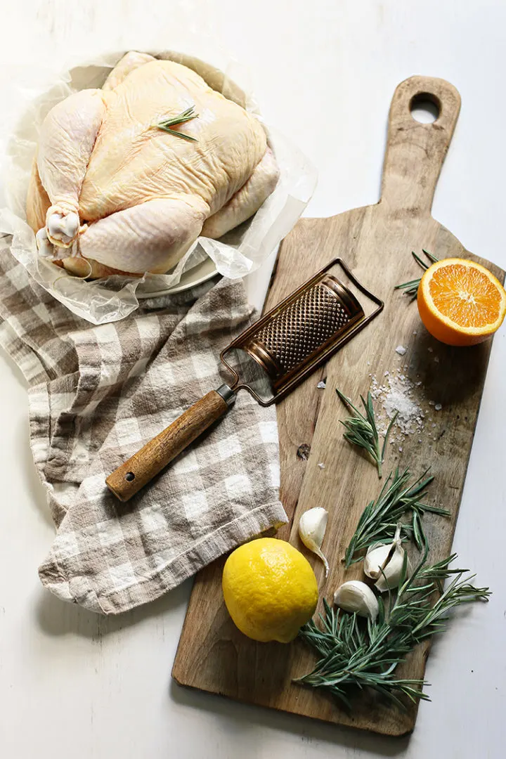photo of ingredients to make roasted rosemary chicken on a wooden cutting board (whole chicken, lemon, oranges, fresh rosemary, garlic, and salt)