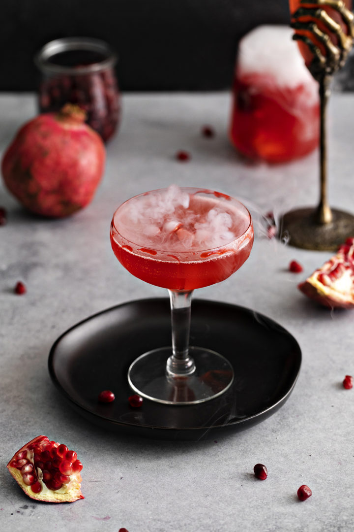 photo of a pomegranate halloween cocktail with dry ice and a fake blood cocktail rim on a black plate on a grey table with surrounded by pomegranates