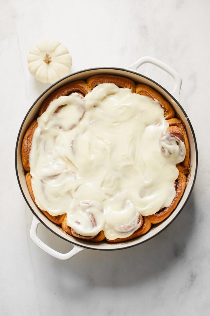 overhead photo of pumpkin cinnamon rolls with cream cheese frosting in a white cast iron pan