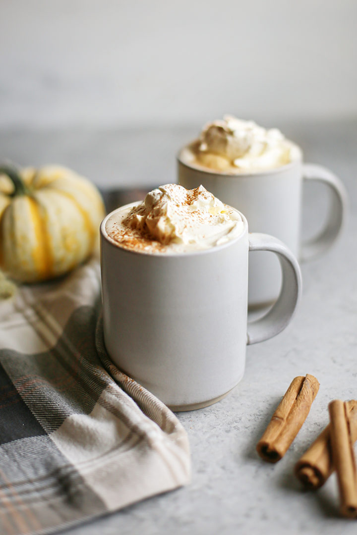 photo of two mugs of pumpkin spice white hot chocolate with cinnamon sticks and a small pumpkin in the background