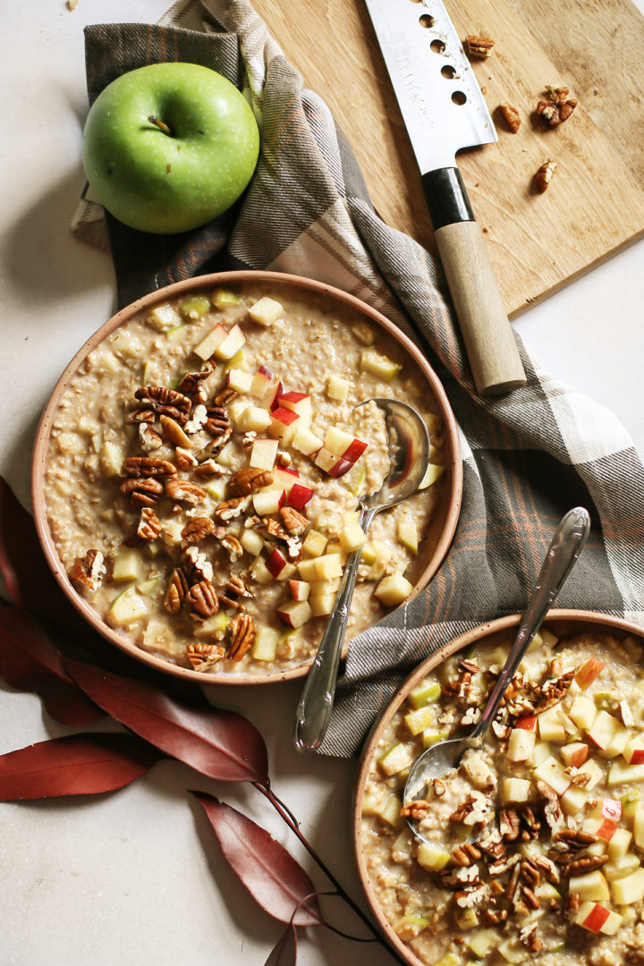 overhead photo of two bowls of apple overnight oats on a table with a plaid napkin, spoons, and red leaves