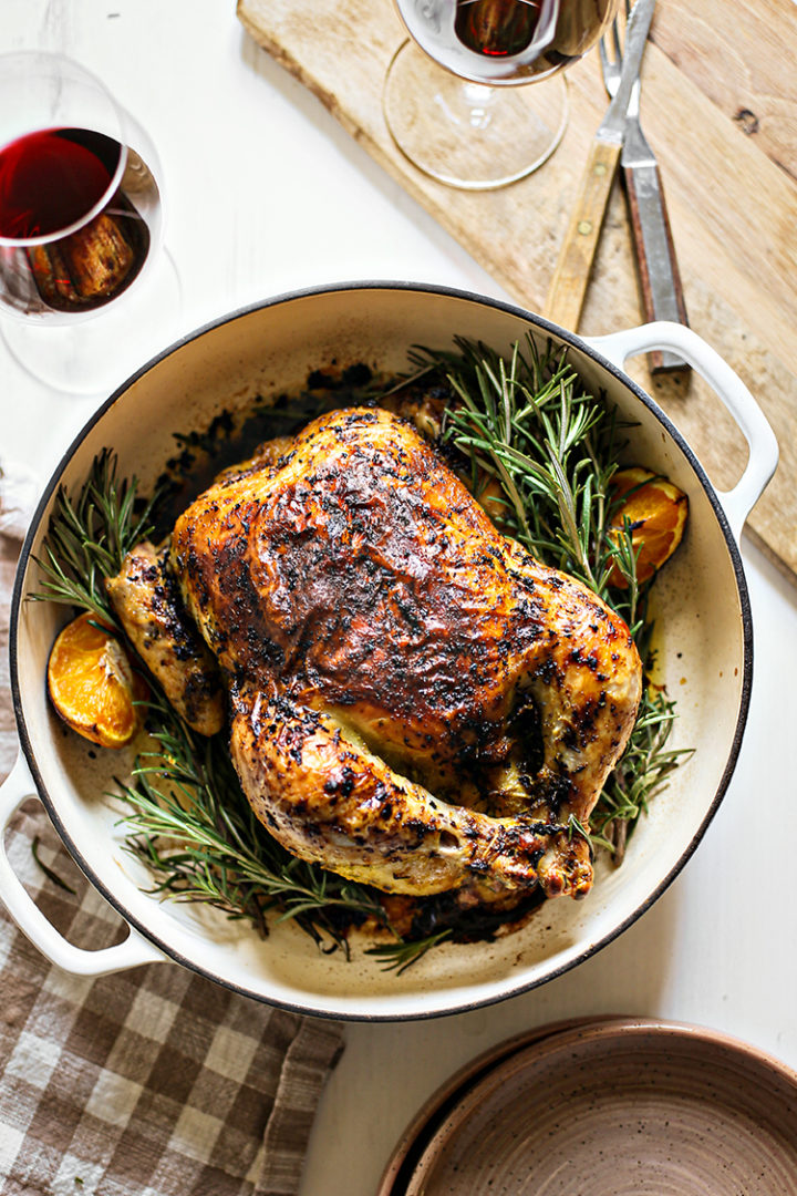 photo of rosemary roasted chicken in a white cast iron roasting pan with fresh rosemary and oranges net to dinner plates, glasses of red wine, and a cutting board