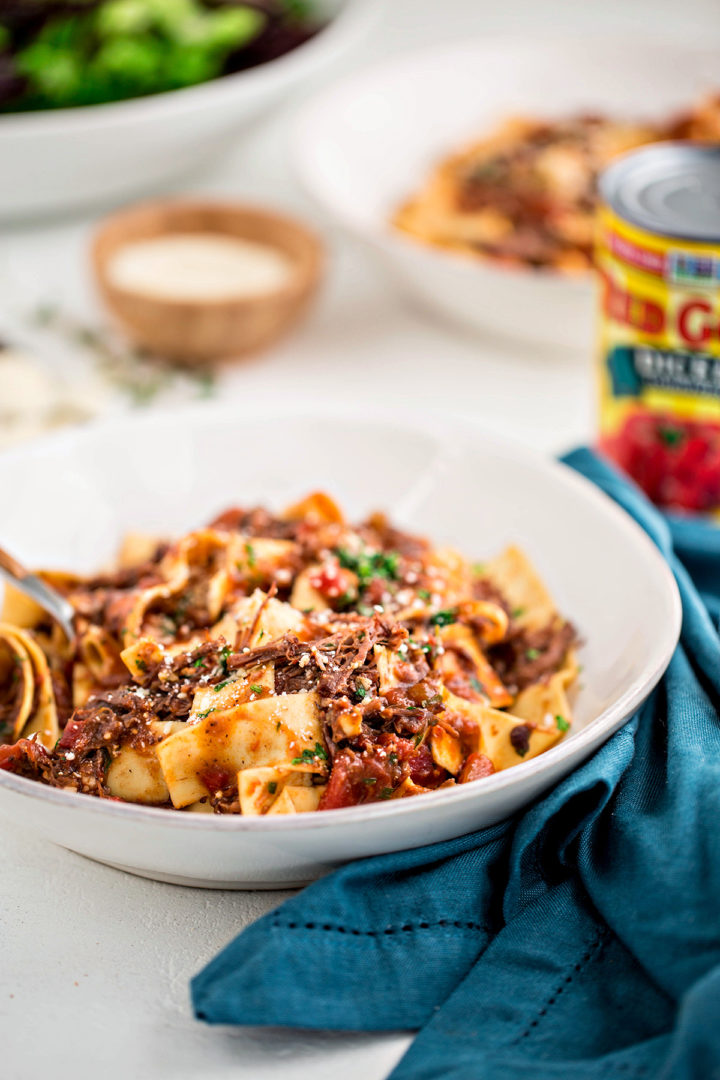 close up photo of a bowl of beef ragu served over pappardelle on a white table