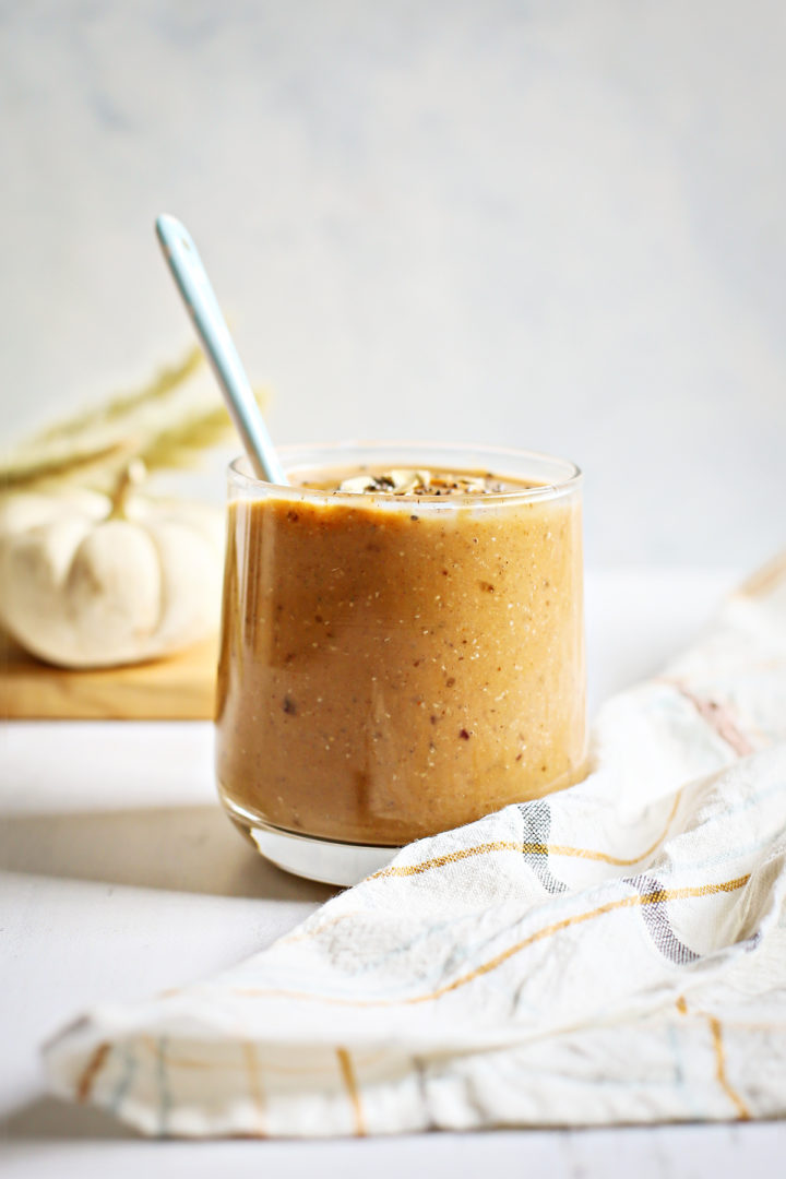 a glass of pumpkin smoothie with a serving spoon on a white wooden table