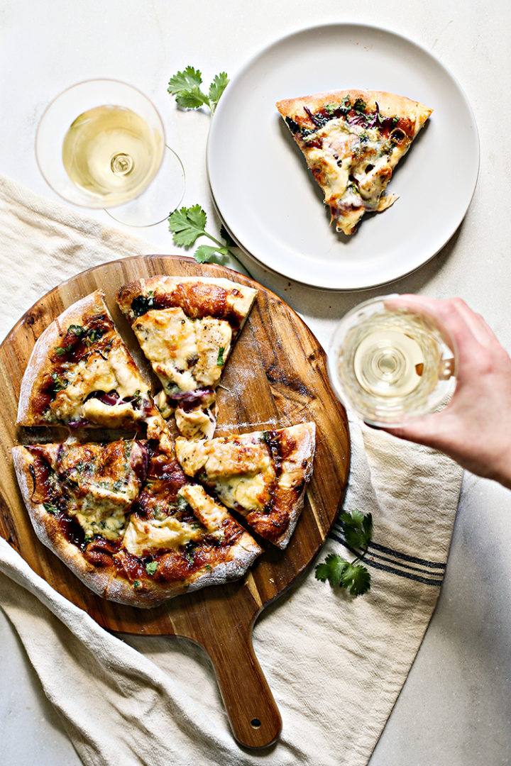 photo of bbq chicken pizza sliced on a wooden pizza peel on a table with glasses of white whine and a slice of pizza on a plate