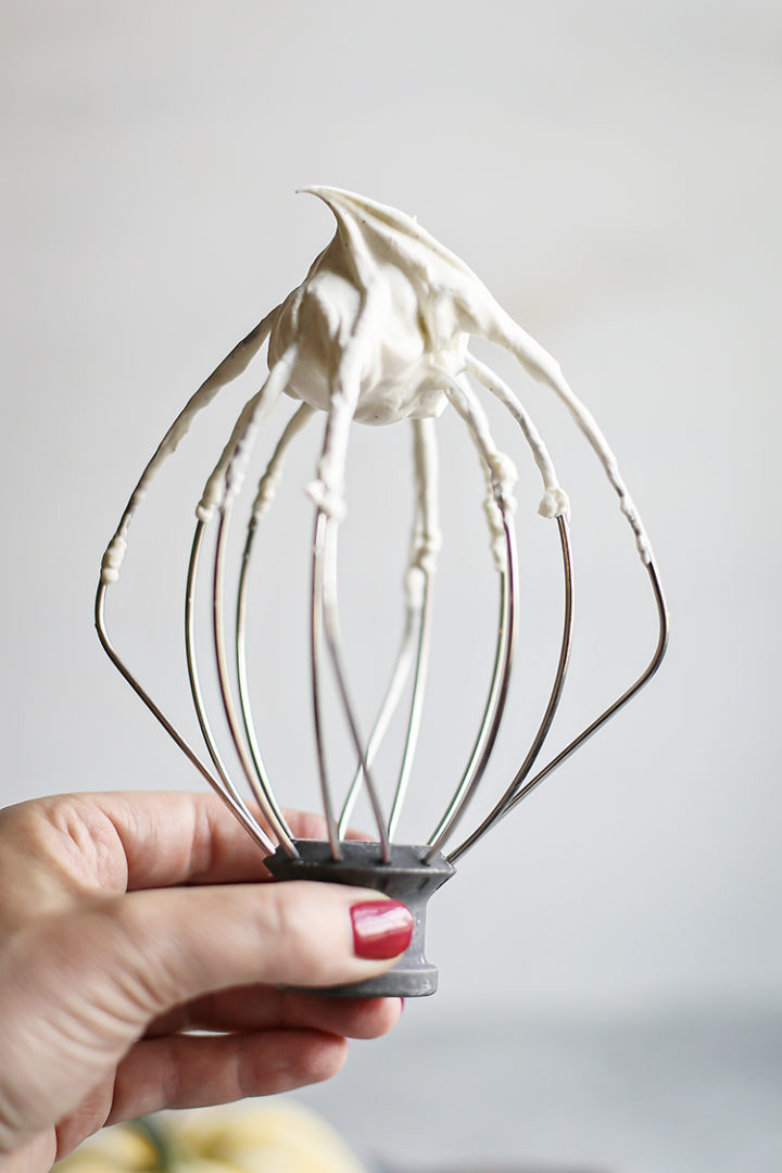 woman holding a wire whisk with vanilla bean whipped cream on it to serve with pumpkin white hot chocolate