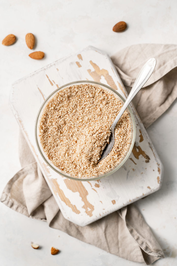 overhead photo of a bowl of almond meal with a spoon in it on a white wooden cutting board