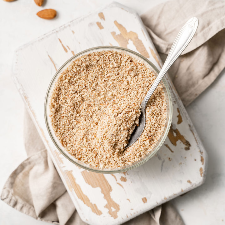 overhead photo of homemade almond meal in a bowl with a spoon on a white wooden cutting board