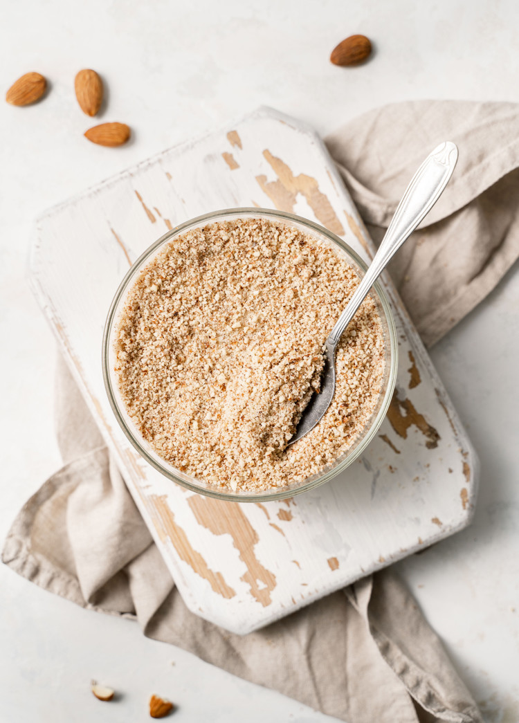 overhead photo of homemade almond meal in a bowl with a spoon on a white wooden cutting board