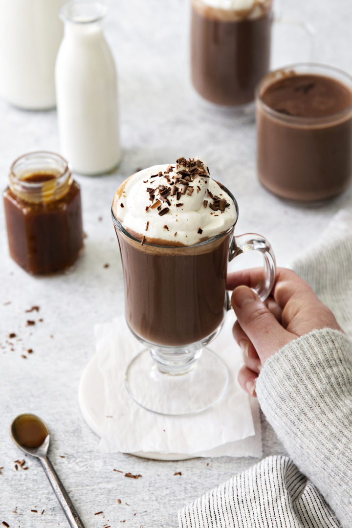 photo of a woman holding a mug of salted caramel hot chocolate with whipped cream and chocolate shavings on top
