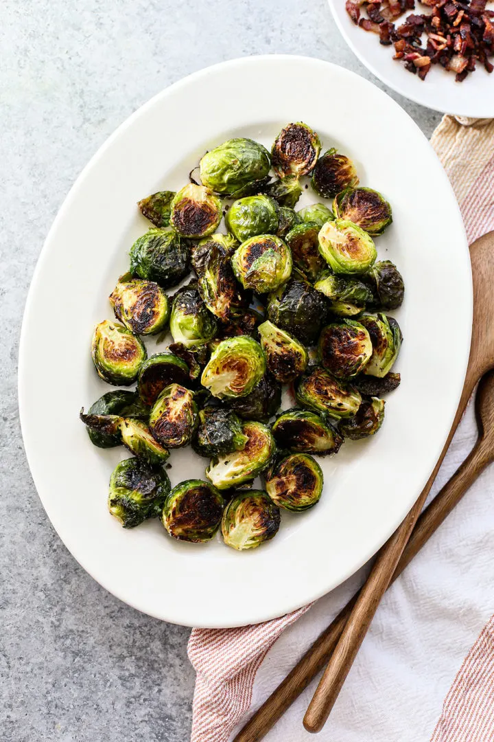 photo of crispy brussels sprouts on a white platter with wooden serving spoons and a bowl of bacon