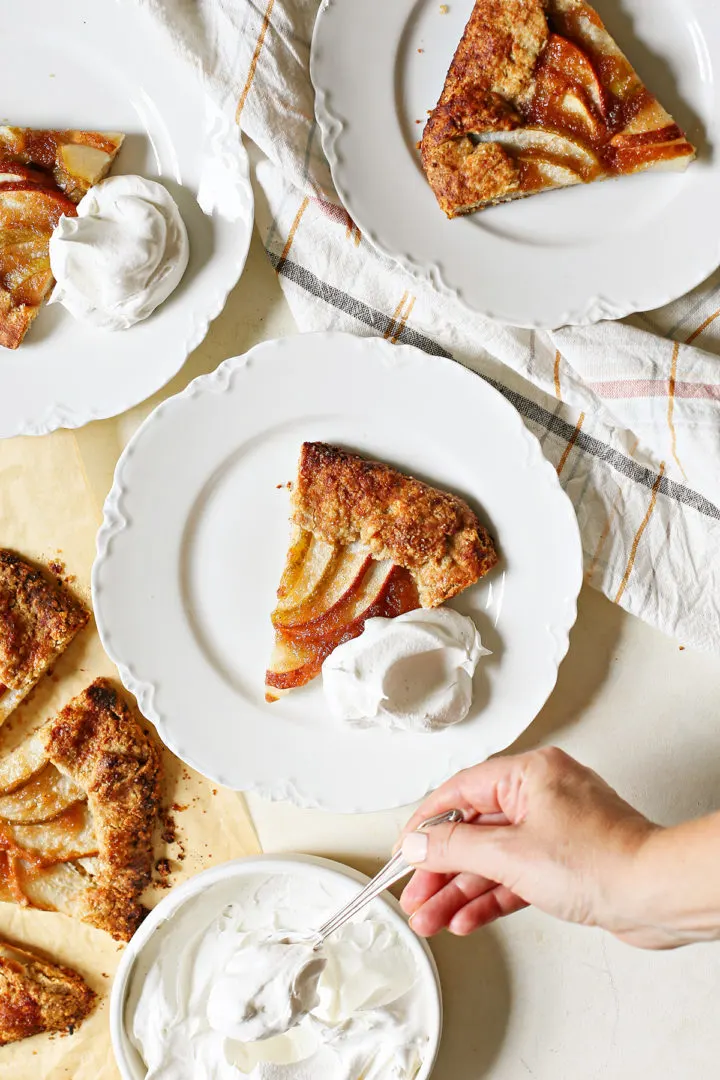 overhead photo of a woman putting whipped cream on plates of pear galette on a white table
