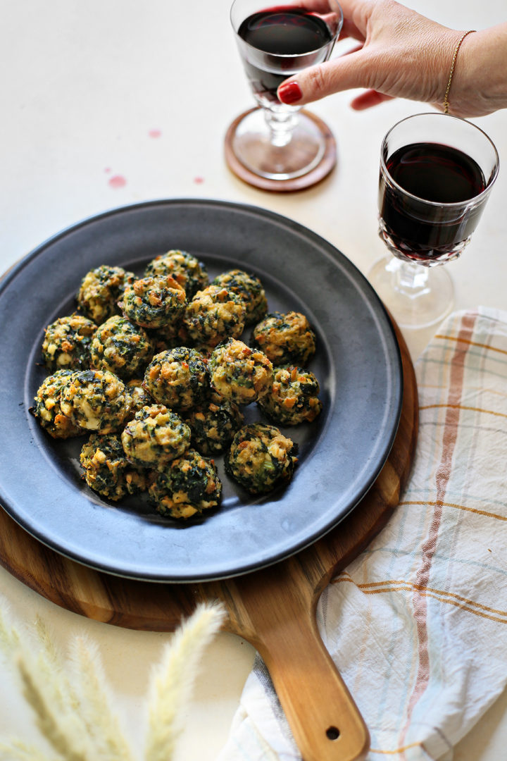 photo of woman picking up a glass of red wine next to a plate of spinach balls