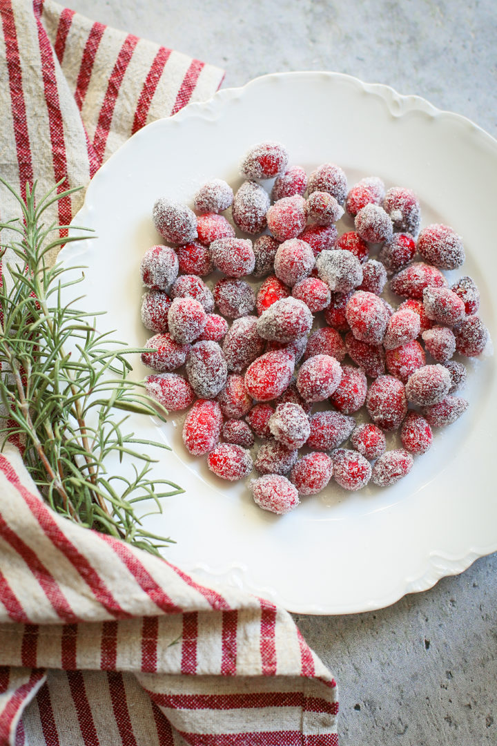 photo of sugared cranberries on a white plate with a sprig of rosemary next to it